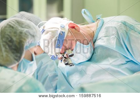 Surgeon and his assistant performing cosmetic surgery in hospital operating room. Doctor in mask wearing surgical loupes. Breast augmentation, enlargement, enhancement