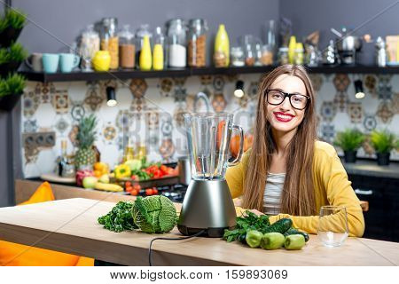 Portrait of young woman sitting on the table with fresh greens and blender at home