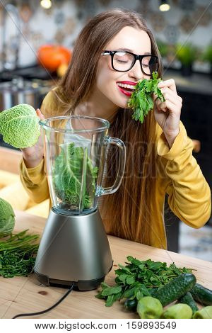 Young smiling woman making smoothie with fresh greens in the blender in kitchen at home. Healthy vegetarian smoothie for weight loss and detox
