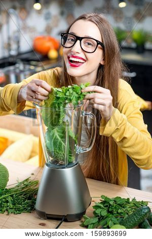 Young smiling woman making smoothie with fresh greens in the blender in kitchen at home. Healthy vegetarian smoothie for weight loss and detox