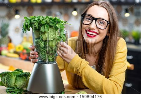 Portrait of young smiling woman with blender full of fresh greens, kiwi and gabbage ready to mix in the kitchen at home. Healthy vegetarian smoothie for weight loss and detox