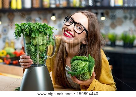 Portrait of young smiling woman with blender full of fresh greens, kiwi and gabbage ready to mix in the kitchen at home. Healthy vegetarian smoothie for weight loss and detox