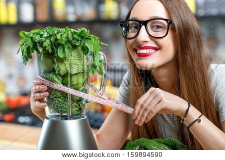Portrait of young smiling woman with blender full of fresh greens, kiwi and gabbage ready to mix in the kitchen at home. Healthy vegetarian smoothie for weight loss and detox