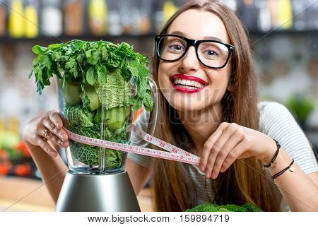 Portrait of young smiling woman with blender full of fresh greens, kiwi and gabbage ready to mix in the kitchen at home. Healthy vegetarian smoothie for weight loss and detox