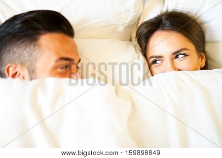 Boyfriend and girlfriend laying in a bed with white sheets - Young and beautiful couple happily looking at each other during bed time - Focus on the girl's face
