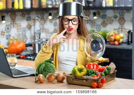 Unhappy woman with cooking pan on the head in the kitchen full of vegetables. Dissatisfaction with the process of cooking