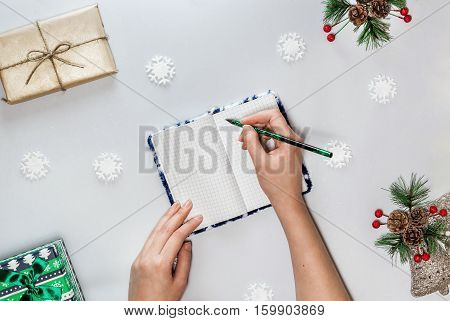 Christmas composition with notebook, female hands with green pencil small green box, snowflakes and spruce branch with red berries and cones. Concept christmas, new year plan. Flat lay. Light gray background