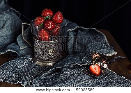 Ripe strawberries in old glass holder on dark rough fabrics background. Renaissance rustic style.