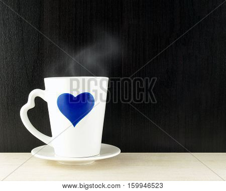 blue heart on side of white ceramic cup with heart shaped handle and saucer on wooden table top and dark wall, hot coffee with steam, relaxing with hot drink in romance mug, close up with copy space