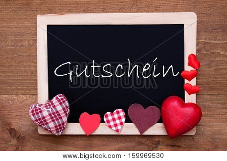 Chalkboard With German Text Gutschein Means Voucher. Red Textile Hearts. Wooden Background With Vintage, Rustic Or Retro Style.