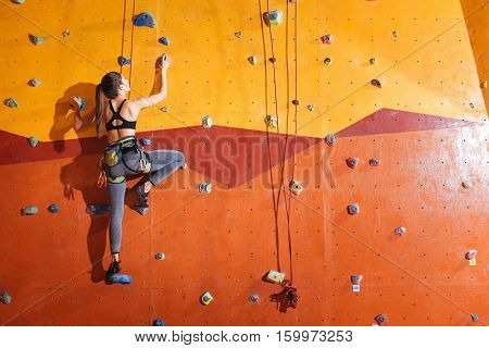 Higher and higher. Attractive young active woman climbing up the wall in gym while training and wearing sportswear and special equipment