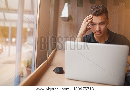 handsome hipster modern man designer working inside cafe at window using laptop. Puzzled young man thinking while sitting at computer with copyspace