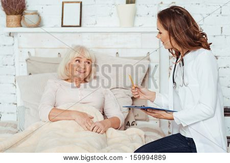 Ready to help you. Pleasant cheerful professional female doctor making notes and examinig her patient at home while aged woman lying in bed