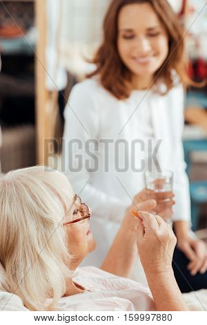 Every day procedure. Pleasant aged woman lying in bed and takign pills while professioanl nurse assisting her