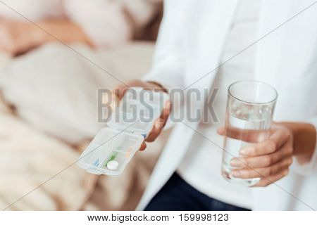 Take care of your health. Close up of pills in hands of professioanl caring nurse holding them and keeping glass of water