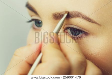 Woman With Blue Eyes Getting Makeup On Brows With Brush. Male Hand Applying Brown Color On Girl Eyeb