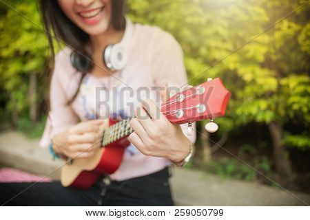 Asian Woman Playing  Song By Red Ukulele. Enjoy Favorite Song On Weekend At Park. Seletive Focus At 