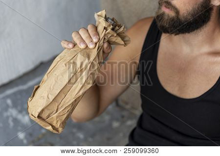 Drunkard Sitting On A Building Rooftop Terrace, Holding A Bottle In A Paper Bag, Drinking. Focus On 
