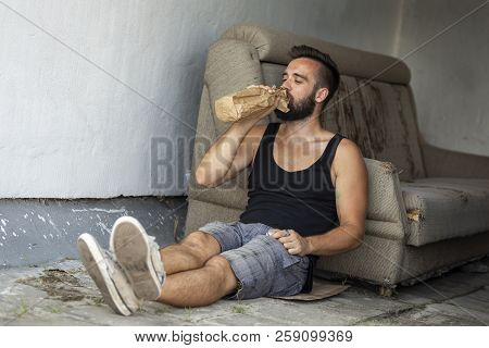 Drunkard Sitting On A Building Rooftop Terrace, Holding A Bottle In A Paper Bag, Drinking