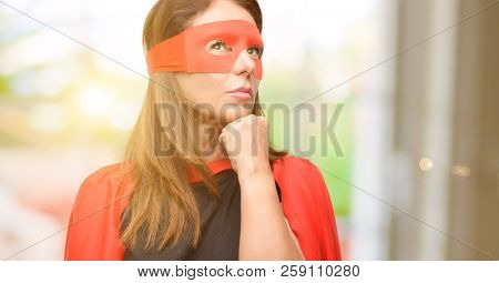 Middle age super hero woman wearing red mask and cape thinking and looking up expressing doubt and wonder