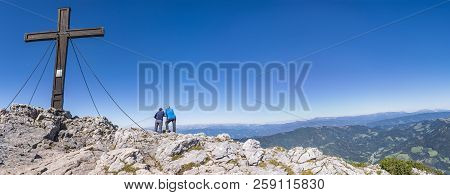 Two Hikers Standing Beside The Cross And Enjoy The Beautiful View From Summit Of Mountain Hochlantsc