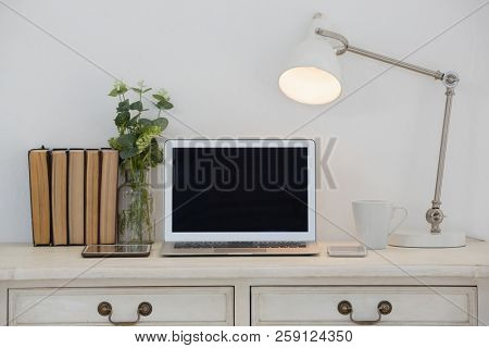 Electronic gadgets, lamp, book, mug, flora and book on table against white wall
