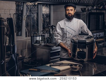 Bearded Cook Dressed In Uniform And Hat Holds Big Multicooker In Kitchen.