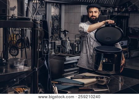 Bearded Cook Dressed In Uniform And Hat Holds Big Multicooker In Kitchen.