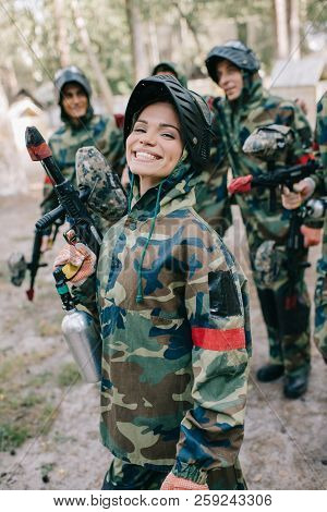 Attractive Smiling Female Paintballer In Uniform Holding Paintball Gun While Her Team Standing Behin