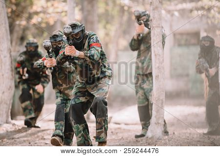 Paintball Team In Uniform And Protective Masks Aiming By Paintball Guns Outdoors