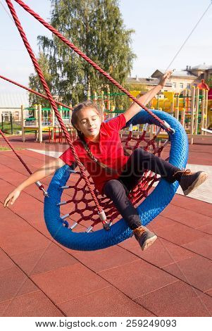 Happy Smiling Child Swinging On A Swing With Closed Eyes And Arms Outstretched At Playground In Summ