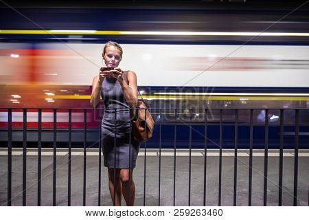 Pretty, young female commuter waiting for her daily train in a modern trainstation, using her cellphone while waiting (color toned image)