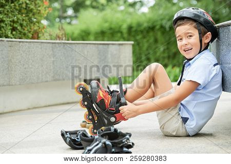 Sweet Asian Boy Smiling And Looking At Camera While Sitting On Ground In Park And Putting On Roller 