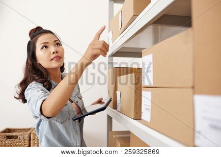 Young Postwoman Using Tablet Pc For Work, She Is Looking For Parcel On The Shelves