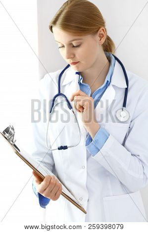 Doctor Woman  Using Clipboard With Medical Record Form While  Standing Near Window In Clinic Or Hosp