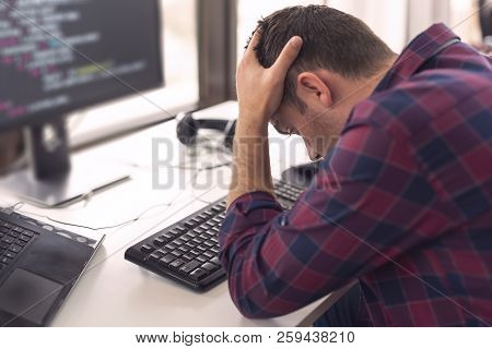 Young Software Developer Sitting At His Desk At Work, Holding Head In Hands, Stressed Out Because Of