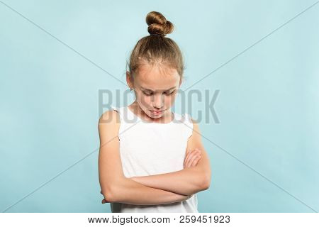 Offended Grumpy Hurt Child With Fold Hands Looking Down. Young Cute Huffy Girl Portrait On Blue Back
