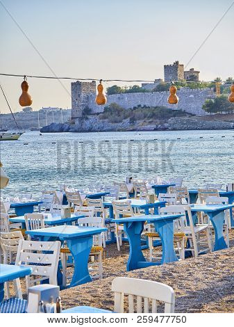 Tables With Chairs Of A Turkish Tavern In A Beach Of Kumbahce Bay With The Castle Of Saint Peter In