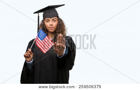 Young hispanic woman wearing graduation uniform holding flag of United States with open hand doing stop sign with serious and confident expression, defense gesture