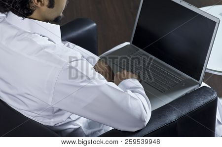 Mideastern Man Using Laptop At Office, With A Cup Of Cofee And Documents On The Side Table