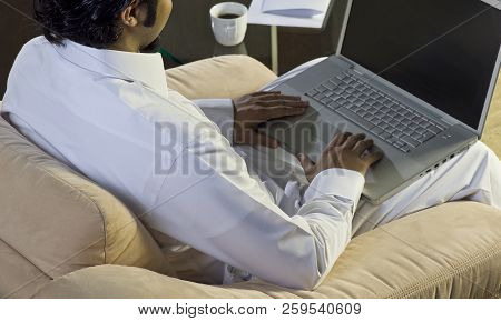 Mideastern Man Using Laptop At Office, With A Cup Of Cofee And Documents On The Side Table
