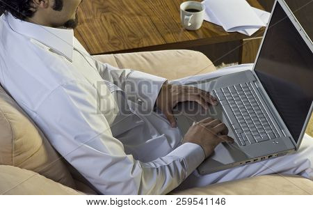 Mideastern Man Using Laptop At Office, With A Cup Of Cofee And Documents On The Side Table