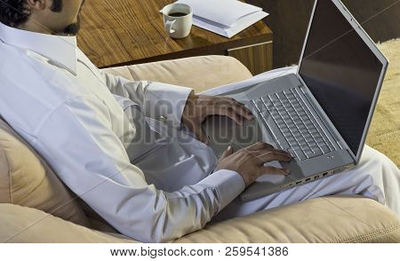 Mideastern Man Using Laptop At Office, With A Cup Of Cofee And Documents On The Side Table