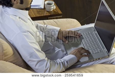 Mideastern Man Using Laptop At Office, With A Cup Of Cofee And Documents On The Side Table