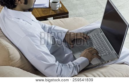 Mideastern Man Using Laptop At Office, With A Cup Of Cofee And Documents On The Side Table