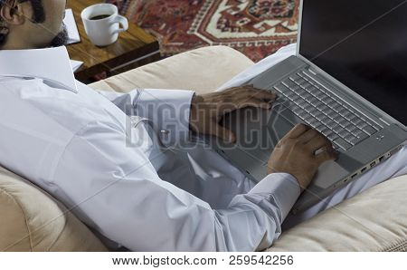 Mideastern Man Using Laptop At Office, With A Cup Of Cofee And Documents On The Side Table