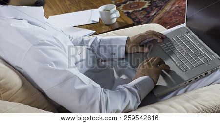 Mideastern Man Using Laptop At Office, With A Cup Of Cofee And Documents On The Side Table