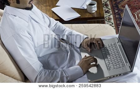 Mideastern Man Using Laptop At Office, With A Cup Of Cofee And Documents On The Side Table