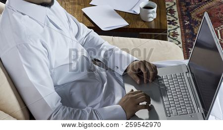 Mideastern Man Using Laptop At Office, With A Cup Of Cofee And Documents On The Side Table