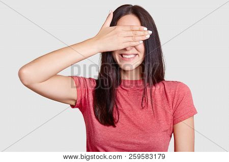 Cheerful Brunette Young European Woman Covers Eyes With Palm, Tries To Hide Her Face, Has Toothy Smi